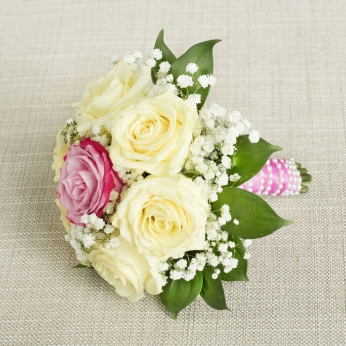 From an overhead, gently slanted angle, this petite bouquet appears ready to star in a Stanmore wedding, glowing gently on a soft, neutral fabric that evokes the refined linens found in local ceremony halls. Five to six creamy, pale yellow roses command the center, their petals exquisitely furled to suggest the beginning of romance or a fresh new day. Slightly off to the left, a vivid fuchsia-pink rose is strategically placed to draw the eye and serve as the bouquet's whimsical focal point-its lively colour capturing the excitement of a celebration on Stanmore's High Street. Sprays of pure white gypsophila are artfully interlaced among the roses, providing texture and airiness that lighten and brighten the design, echoed by glossy, deep green leaves framing the blooms along the bouquet's edge. The handle emerges to the right, wrapped in glossy, fuchsia-pink satin ribbon-its modern flair enhanced by a spiral of dainty, iridescent pearls, which add a subtle sparkle reminiscent of festive evenings in Stanmore. The entire piece sits atop a woven, beige-toned surface, ensuring every flower and detail stands out brilliantly. Perfect for discussions of wedding colour palettes or floral trends in Stanmore, the arrangement projects an unmistakably romantic, joyful, and contemporary aura - ideal for couples and families marking life's milestones in this cherished community.