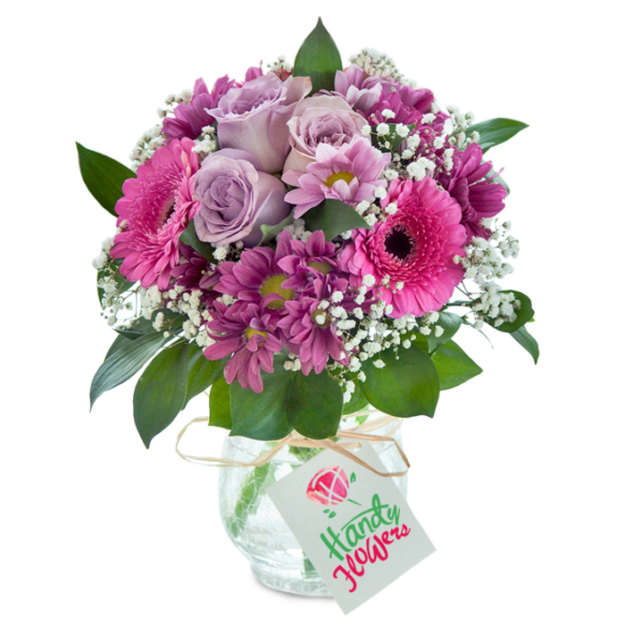 Perfectly capturing the refined energy of Stanmore's landscaped avenues, this exuberant bouquet bursts forth from a clear, textured glass vase, set elegantly against a white background. At its heart, fully open lavender roses unfold gently-each petal delicate and light, giving the impression of early spring in Bernays Gardens. Clusters of dazzling fuchsia Gerbera daisies draw attention instantly, their wide petals and strikingly dark eyes echoing the lively colours that frequent Stanmore's weekend markets. Surrounding these focal blooms, smaller magenta chrysanthemums add intricate texture, their compact layers interspersed with lively yellowish-green central disks that shimmer in sunlight. Ethereal baby's breath is scattered throughout, its clouds of white blossoms lifting the vibrancy of the arrangement and introducing a sense of movement and buoyancy. Deep green foliage frames and supports the blooms, grounding their energy while adding a lush, cultivated finish. The robust glass vase, bearing a subtle cracked-ice pattern, reflects the afternoon light with a twinkling effect, while a rustic wrap of raffia ribbon-tied at the neck-offers a nod to natural simplicity. Completing the ensemble is a pristine white gift tag hanging ready for a custom sentiment. This bouquet evokes heartfelt happiness and refined care, showcasing the artistry that Flowers Stanmore brings to every home, delivered with pride throughout the area for birthdays, thank-yous, or simple daily uplift.
