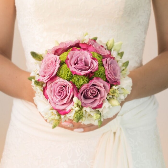Immersed in the gentle ambiance of a Stanmore celebration, this close-up captures the artistry of a hand-tied bridal bouquet set against a stunning white lace gown. The bride's hands, impeccably manicured in light blush-pink with a subtle sparkle, gently cup the rounded composition. At the bouquet's core, large, open roses radiate in tones of lavender, dusty mauve, and touches of vivid fuchsia along the edges-each blossom exuding a sense of romance and thoughtful craftsmanship familiar to any discerning Stanmore bride. Surrounding them, vibrant lime-green button mums introduce lively texture and contrast, reminiscent of the wild grasses and blooms near Stanmore Common. Completing the design, a crown of petite white flowers-probably ethereal freesia or miniature spray roses-creates an impression of freshly gathered blooms, enhanced by small green buds tucked throughout for depth and vibrance. The bridal gown's delicate floral lace motif and refined draping around the waist frame the bouquet beautifully, together suggesting grace and enduring tradition. This artfully constructed bouquet, likely composed by the talented team at Flowers Stanmore, embodies both local flair and classic English romance, offering an elegant inspiration for those planning a wedding or special celebration along Marsh Lane or at historic Stanmore House. The interplay of muted pastels, crisp greens, and soft whites reflects the spirit and charm of Stanmore's community, inviting both joy and reflection.