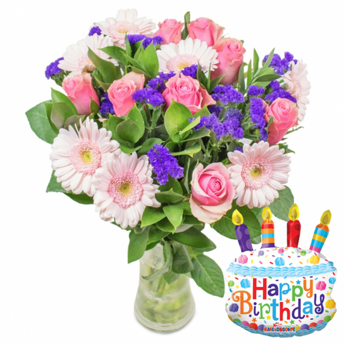 This image captures a celebratory birthday gift with a clear, fresh-focused composition: an abundant bouquet of soft pink roses and pale pink Gerbera daisies is arranged with textural purple filler blooms and glossy green foliage in a transparent glass vase, paired with a whimsical cake-shaped foil balloon. The roses present a range of pinks from the palest blush to a deeper tone, their petals layered and softly ruffled; the gerberas have broad, pale petals edged almost to white and sit forward in the arrangement like bright, joyful moons whose centers glow with yellow-green. Interwoven are clusters of small, vibrant purple blooms - likely statice or limonium - which give the bouquet a crunchy, almost lace-like texture against the smoother rose petals. Tiny white sprays discreetly punctuate the composition, adding fullness and a faint highlight. The vase shows the straight green stems submerged in clear water, reinforcing freshness and careful conditioning. To the right, a white foil birthday cake balloon decorated with blue piping and multicoloured sprinkles draws the eye; four striped candles in purple, red, orange and blue appear alight with yellow flame tips, while Happy Birthday is spelled across the front in playful, colourful letters and a small KALEIDOSCOPE logo rests at the lower edge. Bathed in soft studio light that suggests a calm morning before a celebration, the arrangement feels thoughtful and uplifting - an ideal birthday delivery for someone in Stanmore, perhaps arriving from a local shop near Stanmore Broadway or delivered to a home overlooking Stanmore Park.