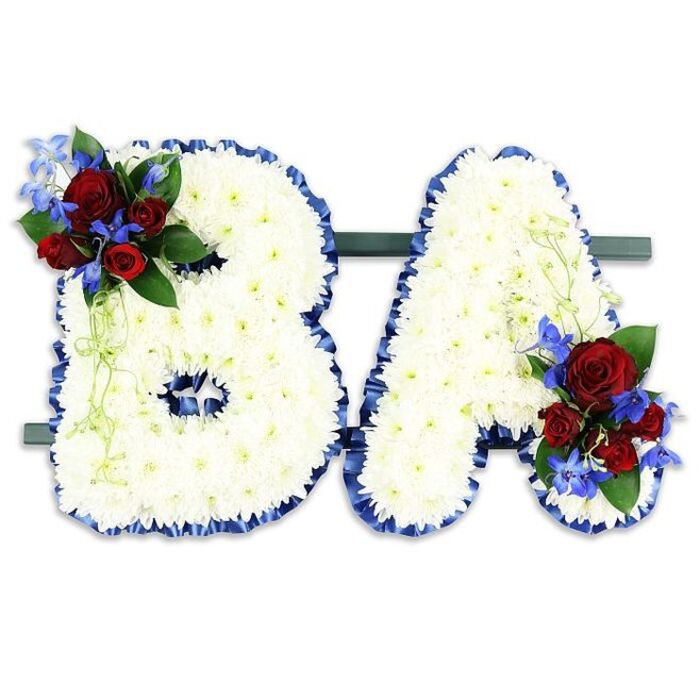 A striking floral tribute crafted to read the initials BA, presented on a dark green horizontal base and photographed against a neutral white backdrop. Each letter is densely covered in tiny, pristine white chrysanthemum blooms arranged so closely that the surface reads like a soft, textured blanket of petals. The strong contours of the B and A are neatly traced with a ruffled royal blue satin ribbon, which both defines the letterforms and introduces a stately, respectful note of colour against the pale flowers.
Thoughtful accenting marks the letters: a rich cluster of deep red roses sits on the upper left curve of the B, their velvety petals opening from tight buds to half-opened blooms, and a matching group rests on the lower right flank of the A. Sprays of intensely blue delphinium-style flowers thread through these clusters, adding vertical punctuation, while pale green trailing foliage and deeper green leaves provide softness and natural movement. The arrangement reads as both architectural and tender-structured initials softened by the tactile layers of florals and greenery. Bathed in soft, even light that lets the white chrysanthemums glow and the red and blue accents hold their depth, this pair of letters would be an appropriate, personalised homage for services in and around Stanmore, from local chapels to memorial gatherings near Canons Park.