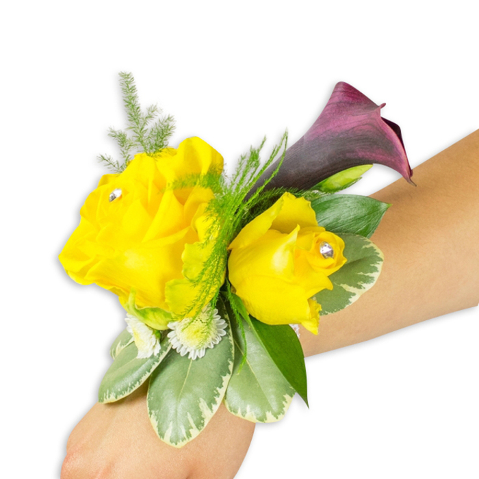 Displayed on a light-to-medium complexion forearm against a clean white backdrop, this wrist corsage balances bold colour with delicate detailing to create a refined accessory for formal occasions. The centrepiece comprises two bright yellow roses: a larger, fully opened bloom to the left and a slightly smaller, more closed rose to the right, both with soft, layered petals that suggest a gentle, classic rose perfume. Each rose is accentuated by a small clear crystal nestled at its heart, adding a poised, sparkling accent. Behind and slightly above the roses, a single deep burgundy-plum calla lily unfurls its sleek trumpet-shaped form, the dark, satiny surface providing striking contrast in hue and line. Fine, feathery asparagus fern threads through and above the flowers, offering lightness and visual lift, while broader variegated leaves with pale edging create a supportive green framework at the base. Tiny white button-like filler flowers - reminiscent of spray chrysanthemums or baby's-breath - tuck into the gaps to increase volume and lend a soft, dotted texture. The corsage sits diagonally across the frame from the lower-left toward the mid-right, demonstrating how the blooms sit when secured around the wrist with a slim ribbon or elastic band that remains mostly hidden. The bright white background highlights each tonal shift - the buttery yellows, the plum-dark calla, and the glossy greens - and the overall aesthetic reads as joyful yet elegant, a thoughtful piece suitable for bridesmaids, prom parties, or a guest of honour arriving at a Stanmore celebration.