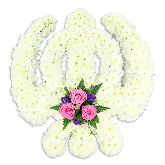 The photograph features a commanding Sikh Khanda floral tribute carefully arranged on a neutral white background, allowing every detail of the design to stand out. The iconic shape is created entirely from dense white chrysanthemums, their rounded faces packed closely together to trace the central double-edged sword, the circular chakkar, and the elegant curves of the crossed kirpans. Because all the structural elements are formed from the same soft white flowers, the design relies on precise craftsmanship and sculpted contours rather than colour contrasts, giving it a sculptural, almost carved appearance. The white blooms glow softly under even lighting, suggesting natural daylight filtering through a Stanmore shopfront window, perhaps along Broadway or close to Stanmore station, where such tributes are lovingly prepared. At the point where the swords cross at the base, a compact spray of accent flowers brings in a touch of colour and tenderness. Three pink roses, their petals layered and velvety, form the heart of this smaller arrangement. Around them cluster several deep purple blooms, rich and saturated, and sprigs of dark green foliage and airy fern leaves that add dimension and natural movement. These colours draw the viewer's attention to the emotional centre of the tribute, representing love and remembrance held within the strength of faith. A soft, diffused shadow beneath the Khanda indicates its depth and three-dimensional form without interrupting the calm purity of the scene. Intended for Sikh funerals, memorials, or commemorative services in the Stanmore area, this floral Khanda expresses profound respect and devotion, blending religious symbolism with the gentle comfort of fresh flowers chosen and arranged with care.