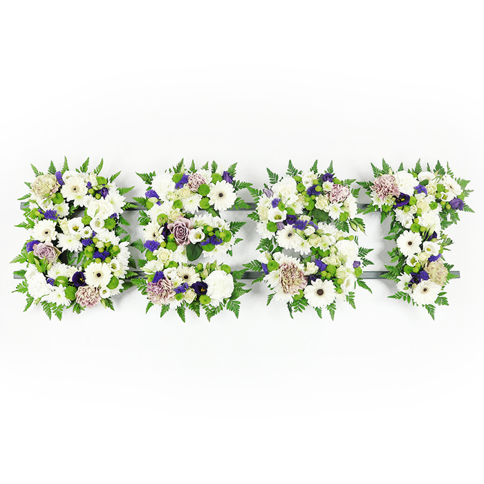 A carefully composed, horizontal floral tribute spells the uppercase word 'BEST' against a clean white backdrop, each letter rendered as a lush cluster of white, soft lavender and verdant green blooms. Prominent are large, pristine white Gerbera daisies with their dark centres, alongside full white carnations and delicate lisianthus or similarly petalled blooms that create layers of texture and softness. Touches of mauve and lavender roses, with their gently cupped petals, and ruffled purple carnations are woven through the blocks of white, introducing a dignified depth and a warm, reverent tone. Along every letter edge, feathery green fern fronds form a soft, natural border that both defines and cushions the shapes, while small green button chrysanthemums are dotted within the clusters to lift the eye and bring a fresh, lively contrast. A dark grey structural base gives each letter its silhouette, slightly visible beneath the blooms and lending a subtle architectural crispness to the overall design. Bathed in gentle daylight the whites read as pure and serene, the purples as thoughtful accents, and the greens as living support, creating a tribute that balances clarity with tender emotion. This style of personalised floral lettering is typically chosen for memorials and funeral services in and around Stanmore - from intimate wakes near Stanmore tube to commemorative gatherings close to Stanmore Common - where families ask for arrangements that honour memory with quiet beauty and a hand-crafted sensibility.