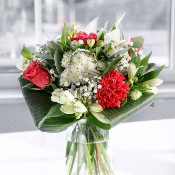 This image shows an elegant, contemporary bouquet presented in a tall, cylindrical glass vase, where the waterline reveals an orderly bundle of fresh green stems trimmed with care. Above this clear base, the arrangement blooms into a rich tapestry of red, white, and green, composed with the balance and precision of an experienced florist. Dominating the design are sumptuous deep red roses, their velvety petals gently opening to form perfect spirals that evoke romance and enduring love. At the front, a large ruffled crimson carnation draws the eye, its densely layered petals providing a striking textural contrast to the smoother rose petals nearby. Threaded throughout the red flowers are serene white blossoms: dainty alstroemeria with slender petals and fine speckles, round white chrysanthemums bringing softness, and several tightly closed white lily buds promising elegant flowers in the days to come. Tiny clusters of gypsophila, or baby's breath, are sprinkled like stars through the composition, adding lightness and a whisper of dreamy charm. Framing all of this colour is an abundance of lush greenery. Broad, dark green leaves, polished to a glossy sheen, are wrapped and curled in a structured collar around the base, while slimmer pointed foliage and eucalyptus-like sprigs weave upward to add movement and varied tones of green. The bouquet rests on a spotless white surface against a softly blurred, bright backdrop that feels like natural daylight filtering through a Stanmore window, perhaps above a shopfront or a quiet home near Canons Park. The atmosphere is fresh and uplifting, making this arrangement ideal as a dining table centrepiece, a surprise delivery for a special birthday, or a thoughtful gift to bring warmth and joy into a Stanmore home, office, or local celebration.