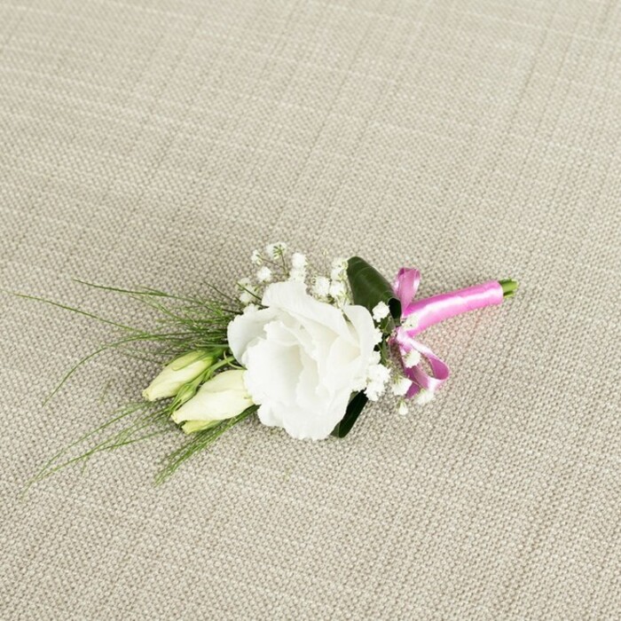 A small, elegant boutonnière is arranged with meticulous care on a light beige, linen-like fabric that shows a fine, grid-like weave, the neutral background recalling the relaxed, natural charm of Stanmore's garden-lined avenues. The arrangement is positioned on a diagonal, with its bloom cluster towards the upper right and the stems trailing to the lower left, creating a gentle sense of movement. Dominating the centre is a fully open white lisianthus, its petals softly ruffled at the edges and layered densely toward the middle, giving it a plush, almost rose-like appearance. Each petal catches the soft, even lighting, revealing subtle shadows and creamy undertones within the white. Just beneath and to the left of this main flower, two elongated lisianthus buds sit close together, creamy and pale green with slightly pointed tips, their closed forms suggesting quiet potential and the unfolding of future memories. Interspersed among the lisianthus are delicate sprays of white gypsophila, tiny round blossoms clustered on fine stems, adding a cloud-like lightness and an ethereal backdrop to the more substantial flower shapes. Slender, grass-like leaves arc outwards from behind, extending leftward and downward like graceful brushstrokes of deep green, while a single broader leaf of a darker shade nestles close behind the open bloom, grounding the design. The stems are neatly trimmed and bound near the bottom right with a bright fuchsia satin ribbon, glossy and smooth, tied into a tidy, cheerful bow that injects a lively splash of colour. Crafted in the style favoured by many couples marrying around Stanmore, this timeless, white-and-green boutonnière with its joyful pink accent feels perfectly suited to a groom's suit, a prom tuxedo, or a special guest dressing up for a celebration near Belmont Circle or Stanmore Broadway.