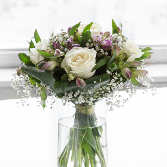 This thoughtfully arranged sympathy bouquet appears as if placed on a polished table in a Stanmore home, catching the gentle midday light that streams in from a nearby garden. Dominating the arrangement are creamy white roses, each bloom full and rounded, with velvety petals that unfurl in balanced layers, forming a focal point of quiet grace and tenderness. Surrounding them, alstroemeria blossoms offer subtle tones of purple and lavender, sometimes tinged with the faintest wash of green at the throat. Some flowers are just starting to open, while others curve gracefully outward, adding movement and complexity to the design. Fine clusters of white baby's breath are woven throughout, like a veil of tiny stars, softening the edges of the roses and alstroemeria and filling the spaces between them with airy lightness. The bouquet is cradled by an abundance of rich green foliage-broad, smooth leaves and slender stems that create depth and contrast, reminiscent of the leafy paths around Stanmore Common. All of this is held securely in a clear cylindrical glass vase, its water fresh and transparent, allowing the viewer to see the straight, carefully trimmed stems aligned within. The background is a softly diffused white, suggesting daylight that has been gently filtered, which makes the whites seem purer and the lavenders more soothing. Together, the elements create a sense of stillness and reflection, making the bouquet particularly fitting for moments of mourning or remembrance. Whether delivered to a residence off The Broadway or to a quiet corner near Stanmore Golf Club, this arrangement offers a subtle yet meaningful expression of support, compassion, and enduring respect.