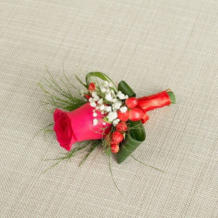 A meticulously crafted floral boutonniere rests at a subtle diagonal on a light beige, textured fabric, the weave reminiscent of natural linen often chosen for elegant wedding styling in Stanmore's intimate venues. The composition moves visually from the lower left to the upper right, guided by the slim, wrapped stem and the gentle sweep of greenery. At its centre, a single striking fuchsia rose becomes the focus, its petals richly coloured and densely layered, forming a tight, velvety spiral that opens outwards into a soft cup, full of depth and freshness. Around the rose, dainty sprays of white baby's breath nestle closely, each tiny blossom forming part of a mist-like cluster that lightens the strong pink and adds a romantic, almost nostalgic softness, as if captured in quiet morning light before guests arrive near Stanmore Country Park. Interspersed among these white florets, several bold red hypericum berries are positioned with care, their glossy surfaces catching the light and their round forms offering a playful contrast to the rose's curves. Fine, feathery strands of fern-like greenery extend gently beyond the main cluster, introducing movement and a touch of wild garden charm, while broader, darker green leaves are artfully curled and layered beneath, providing a sense of structure and framing that reflects a florist's confident technique. The base of the arrangement is tightly bound with a lustrous red satin ribbon, its sheen smooth and reflective, wrapped several times around the stems and finished in a small, secure knot that feels both practical and decorative. Photographed from a slightly elevated angle, every element-petal, berry, leaf, and ribbon-stands out crisply, creating a refined yet approachable piece suited to a groom, groomsman, or prom guest stepping out around Church Road or Stanmore Broadway with understated elegance.