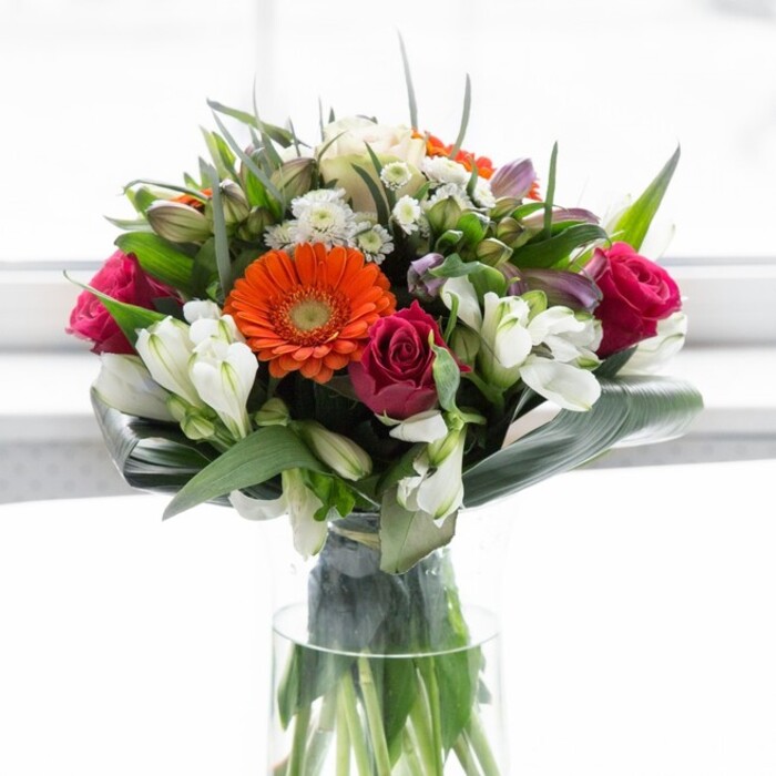 This exquisite floral composition, nestled in a slender glass vase, reflects the refined yet lively spirit of Stanmore and its leafy, peaceful avenues. Creamy peach and vivid orange gerbera daisies cluster at the heart, their soft petals and bold centers reminiscent of festive summer days spent in Canons Park. Intertwined roses in deep magenta bring luxurious drama, while ivory-hued alstroemeria and petite white roses add a delicate, layered freshness throughout the arrangement. Subtle hints of blush pink and the softest touches of airy filler flowers further elevate the bouquet's complexity, much like early morning blooms in a local garden. The entire ensemble is grounded by striking, broad aspidistra leaves carved and curved around the base, creating a protective, sculptural collar that frames the arrangement with organic tranquility. Smaller leaves and fresh greenery add movement, filling spaces between blooms akin to hedgerows lining Stanmore's quieter lanes. Sunlight, diffused and gentle, enhances every colour, lending the bouquet a feeling of hope and renewal. This artful bouquet, perfect for gatherings or sending thoughtful messages, beautifully channels the charm and sophistication of Stanmore's fine floristry tradition.