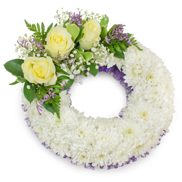 Centered on a pure white background, this dignified circular wreath is crafted from dense, bright-white chrysanthemums arranged into a plush, seamless ring whose open centre suggests calm continuity. A subtle ribbon or mesh of deep lavender-purple peeks through the chrysanthemums along both inner and outer edges, giving the composition a quiet depth and a respectful touch of colour beneath the pristine surface. The upper-left arc of the wreath is punctuated by a thoughtful cluster of three creamy yellow roses; their layered, satin-like petals open gently, offering warmth and a sense of attentive remembrance. Interwoven around the roses are soft sprays of white gypsophila and small, lavender-hued statice blooms that add a floating, delicate texture and a gentle pop of complementary colour. Varied greenery-broad, smooth leaves paired with fine, fern-like foliage-frames the accent grouping and lifts the overall palette with cool green tones. Soft, even lighting emphasizes the wreath's rounded form and casts a faint shadow at its base, lending the photograph subtle dimensionality. Described with the care of an experienced local florist in Stanmore, this arrangement by Flowers Stanmore embodies a respectful tribute suitable for funerals and memorial services near Stanmore station, Stanmore Common, or local churches, conveying serenity and a comforting presence at a time of farewell.