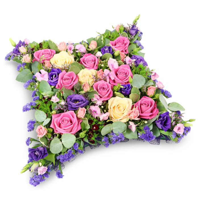 A refined floral pillow tribute is presented in the image, its soft, rectangular cushion shape resting on a stark white background that accentuates the richness of the flowers' colours and textures. This funeral design could easily be imagined placed on a casket or at the front of a chapel in Stanmore, where quiet surroundings and leafy streets lend themselves to thoughtful, elegant goodbyes. The tribute is densely packed, so that petals nearly touch from edge to edge, forming a continuous blanket of blooms. Large roses dominate the composition, offering a sophisticated mix of hues: intense fuchsia roses command attention with their richly pigmented, fully open heads; alongside them, more delicate pastel pink and creamy pale yellow roses soften the overall look, their petals curling gently outward in graceful spirals. Interwoven among these are deep royal purple lisianthus, their multi-layered, slightly frilled petals adding an opulent, velvety texture that anchors the design and gives it depth. Smaller blush pink spray roses punctuate the arrangement, folding into gaps and infusing the piece with a subtle, personal warmth. Fine clusters of lavender and soft violet filler flowers-likely statice or limonium-are threaded through as delicate accents, adding an airy, mist-like halo of colour around the larger blooms. Around the edges and nestled throughout, an assortment of foliage grounds the design: rounded, silvery eucalyptus leaves catch the light with a soft, muted sheen, while darker, slender, fern-like greenery provides contrast and dimension. The lighting is bright yet gentle, creating faint shadows that emphasise the pillow's three-dimensional shape. Overall, the tribute conveys a sense of serenity, comfort, and loving remembrance, making it a particularly suitable floral cushion for commemorating a loved one's life at a funeral, memorial, or graveside tribute in the Stanmore community.