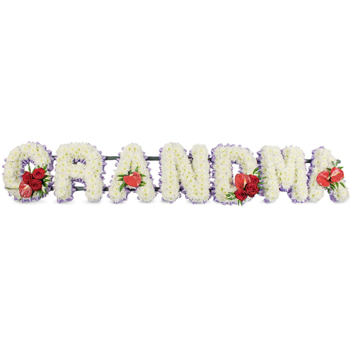 Laid out horizontally against a clean white backdrop, this floral design spells GRANDMA in bold, three-dimensional letters, the sort of tribute often chosen as a focal piece at a funeral in Stanmore or nearby Harrow. Each letter is expertly packed with white cushion chrysanthemums, their small, rounded blooms placed so close together that they form a seamless, cushion-like surface with no gaps. The effect is soft and plush, conveying purity and tenderness, as if the flowers themselves are offering a gentle embrace. Just beneath and around the outer edges of the letters, a fine border of delicate pale lilac ribbon can be glimpsed, tracing every line and curve with a subtle purple edge that gives depth and a quiet, calming hue. Vivid red floral details punctuate the arrangement at key points. On the initial G, a cluster of rich, velvety red roses nestles in the lower curve, joined by a striking red-and-white variegated calla lily whose fluted, sculptural form adds elegance and modern flair. At the first A, a single heart-shaped red anthurium bloom rests neatly at the base, its glossy surface and bright colour standing out boldly against the white. In the centre, the strong lines of the M are softened with another grouping of deep red roses and a second red anthurium, forming a central visual anchor. The final A completes the rhythm with one more red anthurium, ensuring the crimson hearts appear at both midpoint and end. The entire word sits on a dark green supporting frame, likely metal, which lifts and stabilises the letters while remaining mostly hidden. This carefully designed tribute, which a local team like Flowers Stanmore might create for same-day delivery, feels deeply personal and respectful, a fitting way for family members to say goodbye to a cherished grandmother at a church or graveside in the Stanmore area.