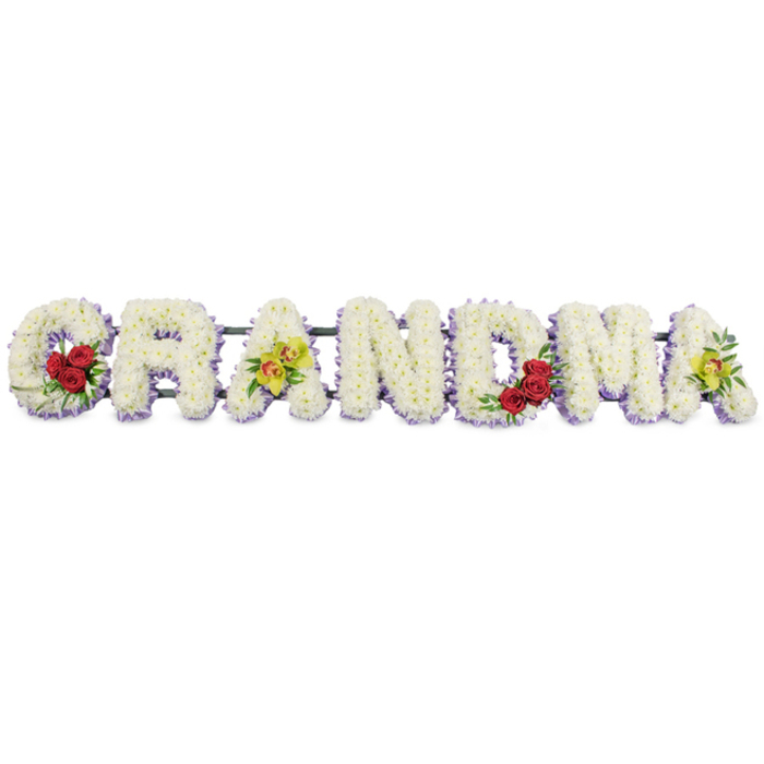 The photograph presents a horizontal floral arrangement spelling out GRANDMA in large capital letters, the type of bespoke tribute often chosen for funerals and memorials around Stanmore. Each letter is completely covered in pristine white chrysanthemums, their small, compact blooms closely arranged so that the flowers form a smooth, almost sculpted surface. The texture is plush yet refined, giving the impression of a soft, continuous blanket of petals rather than individual stems. Along the lower edges and sides of the letters, a subtle border of pale lavender or soft purple flowers runs like a gentle ribbon, outlining the shapes and lending depth, while maintaining the overall calm, understated palette. On the opening G, three rich red roses are gathered at the lower left, their fully open, velvety heads and glossy dark green leaves providing a striking yet tasteful contrast to the white background. The first A features two yellow-green cymbidium orchids resting along its crossbar, their waxy petals and intricate reddish-brown markings adding a note of modern elegance that might evoke a grandmother's unique style or favourite bloom. The central letters R, N, and D are left unembellished beyond their white and lavender flowers, creating a clear and legible centre to the word. Toward the right side, the M repeats the tender red rose detail with another trio nestled into its lower right section, visually balancing the composition. The final A mirrors its counterpart with two more chartreuse orchids, completing the rhythm of the design. All seven letters are mounted on a slender dark green or black metal framework that supports the tribute as a single, stable piece, ideal for resting on a stand at a Stanmore church, funeral director's chapel of rest, or graveside ceremony. The arrangement conveys deep love and respect, offering a focal point around which family and friends can gather to remember a much-loved grandmother.
