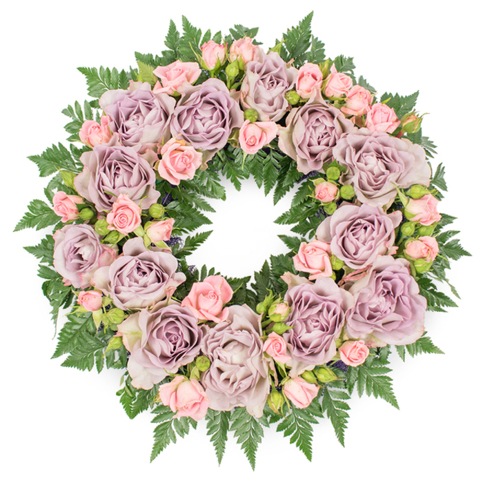 Against a bright, uncluttered white backdrop, a circular sympathy wreath is photographed from directly overhead, allowing every detail of the floral craftsmanship to be seen clearly. The wreath is fully covered with flowers and foliage, forming a lush, continuous ring that feels both comforting and refined, in keeping with the quiet character of Stanmore. Large roses in sophisticated shades of dusty lavender, antique mauve, and soft grey-purple form the primary structure of the design. These roses are fully opened, with numerous ruffled petals that layer one over another, creating soft curves and shadowy centres. Threaded around and between them are clusters of smaller blush pink spray roses, some tightly closed, others just beginning to open, lending a gentle warmth and a more delicate scale to the composition. Beneath and around the roses, a substantial base of dark green fern fronds, likely leatherleaf or sword fern, extends outward in a feathery halo. The deep green foliage frames the pastel blooms, offers contrast, and adds a slight sense of movement with its fine, jagged edges. Subtle darker filler leaves and tiny buds nestle between the main blossoms, contributing nuance and depth without disturbing the overall harmony. The lighting appears soft and even, revealing true colours and the velvety texture of the petals without any harsh glare. The finished impact is one of quiet reverence and comfort, making this wreath an ideal tribute for funerals, memorial services, or commemorative gatherings in and around Stanmore. It is easy to envision it delivered by Flowers Stanmore to a local church, crematorium, or family home as a gentle, dignified expression of condolence, remembrance, and appreciation for a life well lived.
