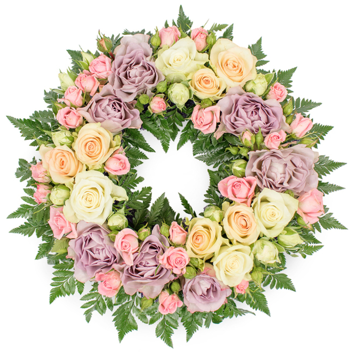 The image captures a lush, circular floral wreath, crafted as a funeral or memorial tribute and placed against a bright white background so that its colours and textures stand out in clear relief. The wreath is richly filled with roses, creating a soft, pastel ring of blooms resting on a bed of dark greenery. Large, fully opened roses in creamy white and subtle pale peach are the most prominent, their petals layered in concentric spirals that give a sense of generosity and comfort. Between them, clusters of smaller blush pink spray roses and tightly closed pale green rosebuds provide intricate detail, making the design feel carefully hand-worked and thoughtfully constructed. Throughout the wreath, you can see distinctive dusty lavender and muted mauve roses, which lend a cool, nostalgic note that beautifully balances the warmer shades. The flowers sit atop finely textured dark green fern fronds and similar foliage, whose feathery edges and rich tones frame the blossoms and add visual depth. The circle is smooth and continuous, without gaps, symbolising eternity, unity, and the enduring nature of love even after someone has passed. The atmosphere is serene and respectful, well suited to accompany a farewell in Stanmore, whether used for a service close to Stanmore Hill or brought to a memorial gathering near the open spaces of Bentley Priory. Arranged in the style favoured by local Stanmore florists, this wreath speaks quietly of remembrance, solace, and the wish to honour a life with gentle beauty rather than grand gesture.