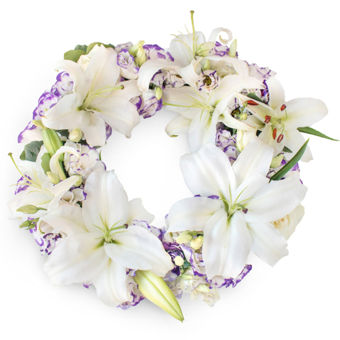 Against a bright, uncluttered white background, this circular funeral wreath appears almost sculptural in its balance of form and colour. The ring is richly covered with large white Oriental lilies, their petals thick, smooth, and slightly curved, giving each bloom a gentle, open-hearted expression. Many lilies face outward around the circumference, showing off pale green throats and elongated stamens tipped with warm, sienna-toned pollen, while a few remain tightly furled, their buds suggesting the tender fragility of life. Nestled among the lilies are clusters of Lisianthus-style flowers, their layered petals white at the centre yet softly edged with lilac and light purple, adding a romantic, whisper-soft hue throughout the design. Occasional small buds-likely lilies or delicate roses-emerge from between the larger blooms, contributing to the cushiony, luxurious texture. Fine sprigs of dark and mid-green foliage, including eucalyptus-like leaves, peek from beneath the petals, giving the wreath a fresh, natural undercurrent, as if it had been composed with care in a Stanmore flower studio near Canons Park. The overall circular form is smooth and uninterrupted, a powerful visual symbol of eternal connection, remembrance, and spiritual continuity. Illuminated by even, gentle lighting, every curve of petal and leaf is visible, with soft shadows adding depth and a sense of quiet presence. The mood conveyed is one of gentle solemnity and comfort, making this wreath a thoughtful choice for funerals, church services, or graveside tributes in and around Stanmore, offering a refined, peaceful way to say a final goodbye.