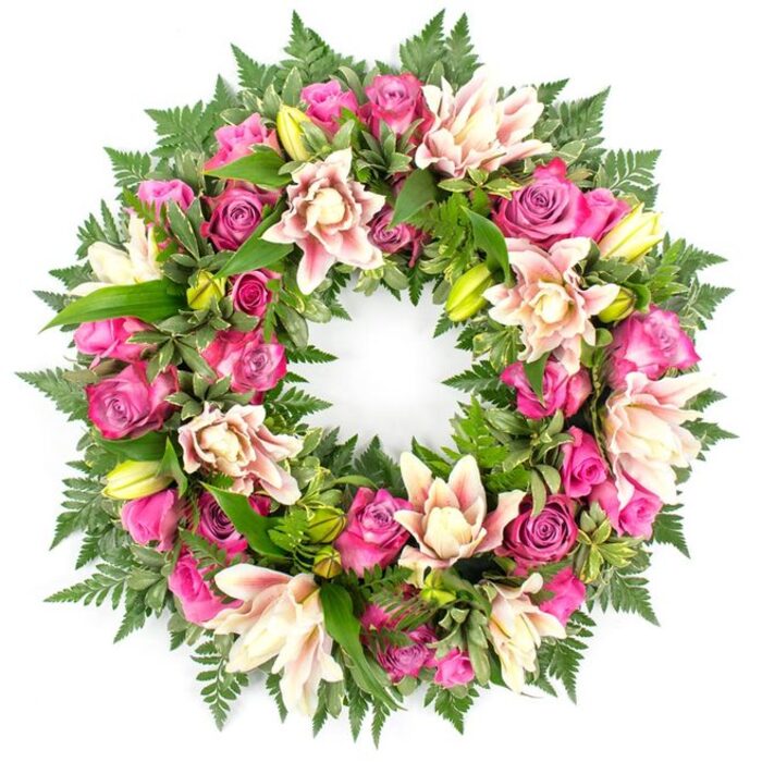 This image shows an exquisitely arranged funeral wreath, set against a clean white backdrop that accentuates every tone and texture. The wreath describes a perfect circle, symbolising continuity, eternity, and an unbroken bond between the departed and those who remember them. The ring is densely packed with flowers, particularly roses in varied shades of pink. Deep magenta and fuchsia roses provide dramatic richness, while softer blush roses offer a gentler, more romantic note. The petals of the roses are thick and velvety, layered so closely that the blooms almost overlap; some are still emerging from tight buds, others are fully opened, revealing complex whorls at their centres. Nestled among the roses are luxurious double lilies, their wide, multi-layered petals in creamy white, faint pink, and a whisper of peach softly reflecting the light. These lilies introduce a sense of airy elegance, while a few unopened buds in vivid green stand tall, adding vertical rhythm and the suggestion of life continuing. The entire arrangement rests on a base of lush foliage. Delicate fern fronds fan out along the perimeter, their fine leaflets creating a soft, lace-like edge. Closer in, glossy leaves of darker green, some perhaps lightly variegated, fill any gaps and support the blooms, creating a rich depth that frames the brighter flowers. The overall impression is carefully balanced: substantial yet gentle, formal yet deeply personal. Crafted by an experienced local florist in Stanmore, this type of wreath could be destined for services at places like St. John's Church, a synagogue off The Broadway, or a quiet gathering near Bentley Priory. The clean white background encourages the viewer to focus solely on the wreath as a symbol of sympathy, gratitude, and heartfelt remembrance.