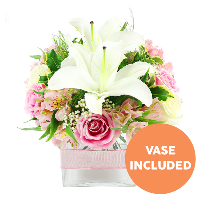 A carefully crafted floral arrangement sits within a clear, square glass vase, photographed in front of a bright white background so that each colour, line, and texture appears vivid and uncluttered. At the heart of the display are two prominent white Oriental lilies, fully opened, their petals gracefully arching outward like sculpted wings. The lilies' centres reveal striking reddish-brown stamens, offering a strong vertical emphasis and a sophisticated contrast against the creamy white. Around these central blooms, a collar of soft pink roses unfolds, with buds in tight spirals alongside more open, cupped flowers whose layered petals give a sense of softness and gentle movement. Interlaced throughout are stems of alstroemeria in blush and white tones, each floret marked with fine yellow accents and scattered speckles, lending an intricate, almost painterly detail to the design. Fine sprigs of white baby's breath stretch lightly between the larger flowers, creating a misty, cloud-like atmosphere that softens transitions and adds a romantic haze. The arrangement is bounded by a rich mix of green foliage-glossy broad leaves, delicate fronds, and varied textures that provide depth and a freshly cut garden feel, reminiscent of a walk through the green spaces around Stanmore Country Park. The stems are cleanly cut to sit within a transparent cube vase, the water line visible and clear, and around the vase's middle a pale pink satin ribbon is wrapped in a straight horizontal band, giving a subtle yet charming decorative accent. In the bottom right corner of the photograph, a circular orange badge with bold white capital text reads VASE INCLUDED, clearly indicating that the container is part of the floral piece. The arrangement has the presence of a small centrepiece, ideal for a Stanmore dining table, sideboard, or bedside, conveying elegance, freshness, and tender celebration.