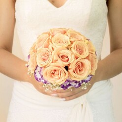 This image offers an intimate, midsection view of a bride holding her bouquet as if it were a cherished secret, the scene likely unfolding in a quiet Stanmore venue bathed in gentle, diffused light. Her white wedding gown, tinged with a warm ivory glow, features fine lace detailing along the bodice and a soft V-neckline that draws the eye toward the floral artistry she carries. The fabric at her waist is subtly gathered, flowing down in graceful folds that provide a serene backdrop for the colours of the bouquet. At the very heart of the design sits a lush dome of roses in soft peach, pale apricot, and blush pink, each bloom tightly formed, the petals curling inward like delicate shells. Their warm tones convey romance and tenderness, as if chosen for a couple exchanging vows in one of Stanmore's quiet churches or garden venues. Beneath this central cluster, a dramatic ring of ruffled flowers-likely lisianthus-wraps the bouquet in a halo of colour, the petals deep purple along the edges before melting into white at the centers. This contrast gives the arrangement depth and a contemporary twist without losing its classic bridal feel. Tiny white flowers, similar to gypsophila, are woven gently around the base, adding a light, airy texture, like a veil of floral lace. The bride's nails are neatly manicured in a soft neutral shade, with a touch of shimmer on one thumb that catches the light. The composition feels serene and thoughtfully crafted, a bouquet that would be lovingly prepared by a skilled Stanmore florist for wedding photographs meant to be treasured for decades.