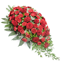 This image shows a refined and dignified red funeral spray presented against a clean white background, so the colours and forms stand out with calm clarity. The design follows an elongated teardrop line, broader and more rounded at the upper left, where the flowers are gathered in their greatest density, then gently tapering to a slim, trailing point at the lower right. Deep red roses are at the centre of the composition, fully opened so that each velvety petal curls slightly at the edge and layers into the next, creating a sense of depth and softness. Nestled closely among them are smaller red spray carnations and traditional carnations, their many fringed petals adding a more textured, feathery look that lightens the visual weight of the roses and introduces a slightly brighter crimson. Around this heart of red, generous greenery creates a lush backdrop. Broad, dark green leaves form a strong structural base, framing the flowers and adding a sense of fullness, while rounded eucalyptus leaves in a soft, silvery-green weave through the blooms, catching the light in gentle highlights. From the lower right, fine strands of trailing foliage extend like a quiet echo of the main spray, giving an impression of natural movement and grace. The overall effect is one of solemn beauty and composed tenderness, very much in keeping with the type of tribute families in Stanmore might choose for a service at a local church or crematorium. Though the image itself is still, you can almost sense the quiet comfort and respect this arrangement offers to those saying goodbye.