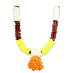 Seen close-up against a stark white background, this U-shaped ceremonial garland reads as a studied composition of colour and texture, imagined for both celebration and reverence. The top sections are built from tight clusters of creamy white petals, their tiny, cushion-like forms conjuring small chrysanthemums and lending a quiet, sacred brightness to the hanging. Below, narrow bands of deep reddish-brown introduce a tactile contrast; these segments look like flattened layered petals or dried bud clusters, suggesting a warmth akin to terracotta or mahogany that anchors the piece. Flowing from those darker intervals, the garland opens into a wide band of vivid golden yellow - a dense, fluffy run of marigold-like elements whose rounded forms create the main U-curve and radiate a convivial, auspicious cheer. A central pendant drops from the meeting point: an exuberant, multi-tiered orange cluster, its ruffled petals arranged to form a generous pom-pom focal point that balances the symmetry. Small green accents are stitched between the colour blocks, read as leaves or ribbon spacers that give structure and gentle visual breathing space. The materials are artificial yet crafted to mimic the fullness and softness of real flowers; under soft lighting the surfaces take on a subtle sheen and would feel silky to the fingertips. Imagined for doorways, temple entrances, or a home puja in Stanmore or nearby Canons Park, this toran evokes ceremony, welcome and the practicality of long-lasting festive decor.