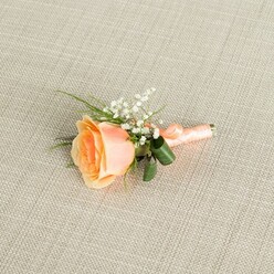 A single peach rose boutonniere is displayed horizontally on a pale beige, linen-textured fabric, the subtle crosshatch of the weave providing a calm, understated backdrop that lets the floral details speak. The rose itself is partially open, its petals curving outward with a soft, velvety fullness, showing an ombré of colour that moves from a rich, warm apricot at the centre to a light, creamy peach along the outer edges. Tucked against the upper right side of the rose is a delicate spray of white baby's breath, each tiny bloom adding a whisper of brightness that contrasts with the rose's smooth, layered petals. Slim green sprigs of foliage radiate gently from behind the flowers, while two broad, darker green leaves are artfully curled around the base of the stems, giving the design a polished, professional finish a local Stanmore florist would be proud of. The stems are tightly gathered and wrapped in a coordinating peach satin ribbon, the fabric smooth and slightly lustrous, ending in a small, neat bow that sits just at the end of the stems. Soft, diffused lighting enhances the natural colours, bringing out the gentle warmth of the rose, the crisp whiteness of the gypsophila, and the cool, calm background tone. The overall impression is of quiet elegance and romance, the kind of boutonniere chosen for a wedding reception near Stanmore Country Park or a prom evening, providing a refined, timeless touch to a suit lapel without overwhelming it.