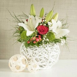 A beautifully balanced floral centerpiece is captured against a softly textured neutral background, the overall effect calm yet celebratory, like a wedding meal unfolding in one of Stanmore's intimate venues. At the centre of the scene stands a spherical white vase, created from interlaced strips that form an open, basket-like weave. It sits on a pure white surface that reflects a gentle light, subtly enhancing the sense of freshness. From this vase, a full arrangement of flowers and foliage fans out in an almost circular silhouette. White lilies dominate the composition, their trumpet-shaped blooms opening in stages; some are fully unfurled, showing delicate markings and prominent yellow stamens, while others remain in green buds that stretch upwards, poised to open over the coming days. Tucked between the lilies are rich red roses, their velvety petals spiralling inward to form deep, inviting centres; one rose stands out near the middle, drawing the eye like the symbolic heart of a wedding celebration. Bright green, rounded flowers-likely pom-pom chrysanthemums or dianthus-are clustered thoughtfully among the larger blooms, contributing both colour contrast and a soft, cushion-like texture. Wisps of airy baby's breath add tiny dots of white that float between the petals, and small clusters of red berries introduce a warm, seasonal accent, suggesting this arrangement could work beautifully for autumn or winter weddings in Stanmore. Lush greenery, including fine, feathery fronds and more substantial leaves, frames the flowers, spilling gently outwards and giving the arrangement a natural, slightly untamed edge despite its overall refinement. On the left side of the frame, two decorative balls crafted from light woven material rest on the same white surface, their patterns echoing the vase and completing a cohesive tablescape. The scene feels polished yet welcoming, the kind of floral design that could sit comfortably on a wedding table in a Stanmore home, local restaurant, or hotel, inviting guests to pause and appreciate its thoughtful details.