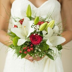 Framed by the elegance of a whiter-than-snow gown, this bouquet in Stanmore captures the spirit of commitment and artistry. The bride's hands curve protectively around a lush design-open white lilies with radiant gold stamens and tightly furled green buds glisten at the focal point, conveying purity and promise. Deep red roses wind through the lilies' embrace, each spiral imbued with stirring passion and enduring love-a signature combination for Stanmore's romantic celebrations. Cheerful clusters of red hypericum berries punctuate the bouquet, their glossy surfaces bouncing with festive energy. Green button-like orbs and airy white Gypsophila create an interplay of texture and movement, echoed in the feathery ferns and polished leaves that cradle the bouquet. The soft light filters through, adding depth and warmth to every petal and leaf. The bouquet's fragrance-roses and lilies, tinged with morning dew-lingers like a whispered blessing. Crafted for a joyful Stanmore wedding, this arrangement is more than decoration: it is a palpable expression of hope and artistry, woven by local florists for same-day celebrations and for memories that last far beyond the aisle.