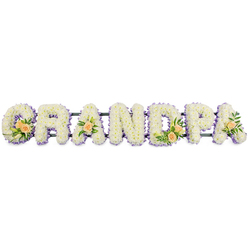 The photograph presents a beautifully composed funeral flower tribute spelling out the word GRANDPA in strong, upright capitals, the sort of personalised piece often delivered by local florists to services around Stanmore and nearby Harrow Weald. Each of the seven letters is generously filled with masses of small, snowy-white chrysanthemums or similar daisy-style blooms, placed so closely that they create the appearance of one soft, continuous surface. The petals overlap like fine fabric, suggesting a sense of comfort and stillness, while the faintest hints of creamy centres add a tiny glow to the design. Around the outer edge of every letter runs a slender border of soft lilac or light lavender flowers, providing a gentle outline that makes each character clearly legible against the stark white background. This muted purple edging deepens the design without overpowering its peaceful tone. Interspersed among the white blooms are carefully positioned accents of peach roses: fuller clusters on the G and D, and single, elegant roses on the first and last A. These roses vary from open blooms to tighter buds, their warm peach petals complemented by fresh green leaves and occasional narrow white lily buds that add a touch of height and movement. The letters are aligned along a discreet, dark horizontal frame which supports the tribute and keeps all seven characters steady and level. Overall, the arrangement conveys tenderness and respect, with the combination of white, lilac, and peach expressing love, remembrance, and gratitude for a grandfather whose presence is still very much felt in the hearts of family gathered in Stanmore.