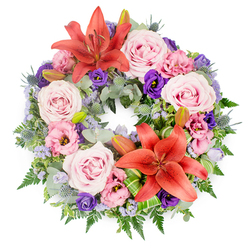 Resting against a clean, white backdrop, this sympathy wreath appears as a complete, unbroken circle of colour, suggesting continuity and everlasting affection. Two striking coral-orange Asiatic lilies, one near the top and one nearer the base of the ring, punctuate the design with warmth and gentle vibrancy. Their open petals curve outward gracefully, revealing rich stamens that offer subtle texture and depth. All around them, clusters of soft pink and lavender-pink roses are nestled closely together, each rose at full bloom with layered, velvety petals forming elegant spirals. Intermixed with these roses are lisianthus blooms, some pale and blush-toned, others a deep, dignified purple, their ruffled petals adding a feeling of softness and quiet luxury. Fine sprigs of lavender-blue filler flowers are scattered throughout, airy and delicate, softening the edges between the larger blooms and creating a light, cloud-like effect. The wreath is supported by a collar of varied green foliage: silvery eucalyptus leaves that add a cool, muted backdrop, slender fern fronds that bring gentle movement, and small blue eryngium thistles, whose spiky forms introduce a subtle architectural note. Tucked within the greenery are several closed lily buds, carefully placed to suggest future blossom and the continuation of memory. Bright green, shaped leaves, possibly ti or aspidistra, curve neatly around the focal lilies, showcasing the florist's skill and adding a contemporary twist to a traditional tribute. Designed in a style that would be at home in Stanmore's local chapels and crematoria, this wreath feels calm and respectful. It would make a fitting floral expression of sympathy for services near Stanmore Country Park or for delivery to bereaved families across Stanmore, carrying with it a message of remembrance and enduring love.