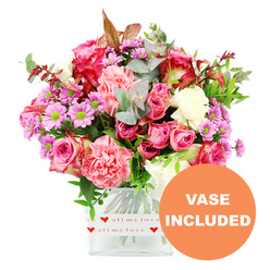 This image shows a generous, hand-crafted bouquet displayed in a square clear glass vase, photographed head-on against a clean white background that echoes the tidy elegance of a Stanmore living room. The arrangement is a celebration of pinks: large, velvety roses open in layers of deep fuchsia and magenta alongside softer blush and pastel shades, giving the bouquet a romantic, storytelling feel. Between them, plump, ruffled carnations in coral-pink and peachy tones form cushions of texture, like soft fabric folded around the roses. Sprinkled throughout are delicate, daisy-style chrysanthemums in lavender and light purple, their vivid lime-green centres providing bright pinpoints of contrast that lift the entire colour scheme. Creamy white blooms lie tucked between the stronger hues, adding a sense of air and spaciousness. The flowers are held together by plentiful greenery: smooth, rounded eucalyptus leaves in soft blue-green, richer dark green foliage, and a few small sprays of reddish-brown leaves that give the design a slightly seasonal, intimate note. The clear glass vase, with its neat, contemporary lines, is wrapped at the base with a white band decorated in a repeat pattern of red hearts and the phrase "all my love" in flowing red script, making the container itself part of the message. In the bottom right corner, an orange circular emblem with bold white capital letters reads "VASE INCLUDED," drawing attention to the ready-to-display nature of the design. The overall impression is of a warm, caring bouquet you might have delivered to a home off Stanmore Hill, an apartment near the station, or a family house close to Canons Park, to celebrate a birthday, anniversary, or simple everyday affection.