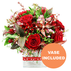 This image shows a sumptuous, love-themed arrangement placed against a pure white backdrop, its reds and greens glowing as though lit by soft daylight. At the center, several large red roses unfurl in different stages of bloom, their velvety petals ranging from tightly furled spirals to fully open, layered rosettes, conveying both drama and tenderness. Echoing their passionate hue, deep crimson accent flowers, likely alstroemeria or a similar variety, nestle between the roses, their slender petals and intricate markings creating movement and subtle pattern. Threaded between these stronger elements are delicate sprays of tiny pale pink and white waxflower-like blossoms, with minuscule cups and buds that resemble a dusting of floral lace, lightening and softening the bouquet. Enveloping the blooms is an array of lush greenery, from broad, smooth leaves to finer, more textured sprigs, which provide fresh contrast and structure. Striking burgundy and reddish-brown leaves, some gently curved and finely veined, extend outward, introducing a note of refined drama that feels well suited to a stylish home near Stanmore station or along The Broadway. The composition forms a full, rounded dome, thoughtfully constructed to appear generous from every angle, reflecting the practiced hand of an experienced florist. All the stems are arranged within a sleek, clear rectangular glass vase, where the straight edges offer a contemporary counterpoint to the natural forms of the flowers. A red ribbon adorned with repeating white heart outlines encircles the upper portion of the vase, quietly reinforcing the romantic message. In the bottom right corner of the image, a circular orange overlay displays the phrase VASE INCLUDED in bold white lettering, reassuring anyone ordering this bouquet for delivery in Stanmore that the gift comes complete, ready to place on a dining table, bedside, or office desk.