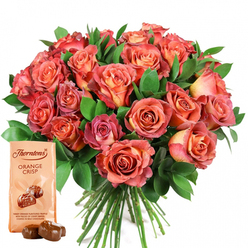 Prepared with artistry befitting the Stanmore community, this thoughtfully curated gift set is a heartfelt tableau of luxury and emotion. A sumptuous bouquet bursts with roses in a harmonious blend-coral, peach, and salmon petals spiraling to pink and cream at the edges, each flower's color softly flowing like the gentle dusk skies over Stanmore Hill. The velvety blooms are held close, their fullness enhanced by interspersed sprigs of polished, dark green foliage, perhaps reminiscent of the lush gardens that flourish along Marsh Lane. Lighting catches the delicate texture of every petal and leaf, suggesting softness to the touch and inviting the fragrance of freshly cut roses. Completing the ensemble, the lower-left corner presents a pale beige Thorntons Orange Crisp chocolate pouch, upright and adorned with signature branding and an irresistible depiction of two chocolate truffles-a promise of crisp orange truffle centers wrapped in milk chocolate. Two of these truffles gleam on the backdrop before the pouch, their deep brown sheen offering a moment of indulgence for any celebration. The entire composition radiates with the possibility of joy-perfect for anniversaries, Mother's Day, birthdays, or any moment deserving a little extra warmth, all delivered across Stanmore with signature local care.