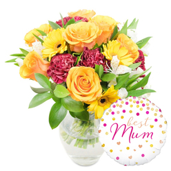 A warm, cheerful bouquet paired with a playful message balloon, arranged and lit to feel like early-afternoon sunshine in a family kitchen. Set against pristine white, the clear glass vase reveals neatly trimmed green stems standing in fresh water, evidence of a recently made arrangement destined for someone special in Stanmore. Dominant peach-orange roses, full and gently unfurling, draw the eye with their soft, petal-rich centres; they give the bouquet a classic, affectionate tone. Interspersed are radiant yellow gerbera daisies whose flat faces and darker centres punctuate the composition with bright optimism. Rich, deep-red or burgundy carnations add a plush, textured counterpoint - their ruffled edges creating shadows and tactile interest - while narrow, speckled white alstroemeria provide small, delicate highlights that lift the palette. The foliage is abundant and varied, from glossy broad leaves that anchor the design to fine, feathery sprigs that add movement and breathe between blooms. Nestled at the base and overlapping the vase, a round white foil balloon carries a scattering of gold and hot-pink polka dots and tiny hearts; across its centre, a lively hot-pink script proclaims "best Mum," a personal touch that turns the arrangement into a gift-ready statement. This composition feels particularly suited to deliveries across Stanmore and nearby Canons Park or to a thoughtful surprise left on a doorstep along Stanmore Hill - fresh, heartening, and unmistakably celebratory.