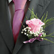 This image offers an intimate, waist-up view of a person dressed in refined formalwear, the camera lingering on the details of a rose boutonniere perfectly pinned to a dark suit lapel. The suit, a deep charcoal or almost-black shade, is sharply tailored, its lapel line clean and structured, hinting that the wearer may be heading to a wedding at a Stanmore church or a black-tie event near Canons Park. Beneath the jacket, a light dress shirt with a subtle gingham or check pattern brings gentle texture, while a vivid fuchsia tie pours down the centre like a bright ribbon of colour. The tie's smooth, satiny surface catches the light, providing a bold accent against the darker cloth. Anchoring the look is the boutonniere, clearly the florist's pride: a single, well-formed pink rose, fully unfurled but still crisp at the petal edges, shifts from a slightly deeper pink at its base to a soft, romantic blush toward the outer curls. Surrounding the rose, clusters of white baby's breath create a cloud-like halo, their tiny star-shaped flowers giving a delicate, lacy effect. Slender strands of fresh green foliage extend upward in soft arcs, adding height and an organic, garden-inspired note that contrasts with the suit's sleek geometry. At the base of the stems, a deep green wrapping is cinched by a narrow mauve or lavender ribbon, tied neatly into a small bow that rests just above the breast pocket. The purple tone of the ribbon harmonises with the pinks in the rose and tie, weaving everything into a cohesive palette that feels affectionate and hopeful. The overall mood is one of tender anticipation, as if the wearer is about to step out into a memorable Stanmore celebration where every detail, down to this single flower, has been chosen with care.