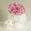 A serene indoor scene shows a rounded bouquet of soft pink roses and white baby's breath resting in a white woven vase, styled as if ready to grace a calm corner of a Stanmore home. Around fifteen to twenty roses are clustered closely together, each bloom opening in generous layers that shift from a pale blush at the centre to a deeper, almost fuchsia pink along the petal edges. The roses are threaded through with sprays of delicate white gypsophila, their tiny, cloud-like blossoms lightening the arrangement and giving it a whispery, romantic softness. The flowers sit in a spherical vase crafted from interlaced white strands that resemble painted wicker or rattan, with an airy, open lattice that lets light pool around the base and cast subtle shadows on the smooth white tabletop. Just to the side of the vase, two decorative orbs made from pale natural fibres lie on the surface, their criss-crossed strips echoing the texture of the container and adding a contemporary, minimalist touch. The whole composition is set against a warm beige wall with a gentle, almost fabric-like texture, softly lit as though by natural daylight filtering through a window on a quiet residential street in Stanmore. The mood is peaceful and refined, suggesting a thoughtful gift for a birthday, anniversary, or Mother's Day, or a graceful floral accent for a living room or hallway near Stanmore Country Park, where soft colour and natural materials bring quiet joy into the space.