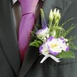 A close-up photograph shows the upper torso of a man dressed in elegant wedding attire, standing in soft, natural light that seems like it could be filtering through a Stanmore church doorway or a garden marquee. His dark charcoal grey suit jacket is sharply tailored, with a clean, structured lapel that frames a pale grey checkered dress shirt beneath. At the centre of the image, a vivid magenta silk tie, expertly knotted, glows with a gentle sheen, adding a modern splash of colour against the muted tones of the fabric. Pinned carefully to the left lapel is a detailed boutonniere that clearly has been crafted by an experienced florist. Two white lisianthus blooms, their ruffled petals edged in a rich, painterly purple, sit at the heart of the arrangement, their yellow centres just visible like tiny sparks of light. Around them, slender white buds reach upwards, still closed with promise, and a single vertical line of tiny deep-purple veronica florets adds height and a touch of wild garden character. Wisps of feathery greenery and fine loops of decorative grass mingle with glossy emerald leaves, giving movement, texture, and a fresh, almost meadow-like feeling. Small hints of delicate white filler flowers, reminiscent of baby's breath, soften the composition. At the base, a neat white satin bow gathers the stems together, suggesting tradition and quiet care. The overall effect is one of poised, joyful anticipation, perfectly suited to a groom or groomsman at a Stanmore wedding, perhaps just moments before stepping into the ceremony to meet the day's celebrations with calm, heartfelt confidence.