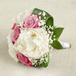 Resting on a softly woven, light beige fabric that resembles natural linen, the bouquet in this image is arranged horizontally, as if placed carefully on a bed or sideboard in a Stanmore home before being picked up for the walk down the aisle. Dominating the centre is a sumptuous creamy white peony, its bloom generously open, with layer upon layer of gently frilled petals forming a rounded, cloud-like heart. Encircling this focal flower are several fuchsia-pink roses, their rich colour lending warmth and a sense of classic romance, while their tightly spiralled petals suggest that they have been freshly arranged and are just beginning to unfurl. Interspersed throughout the composition are fine sprays of white gypsophila, also known as baby's breath, with tiny clustered blossoms that add a lace-like delicacy, filling small gaps and softening the transitions between the main blooms. Framing the entire cluster, deep green leaves sweep outward in smooth arcs, their surface slightly glossy, providing contrast, structure, and a feeling of garden vitality that recalls early morning walks through Stanmore Country Park. The stems are drawn neatly together and wrapped with a smooth band of white satin ribbon, wound with practiced precision to create a sleek, functional handle ideal for a bride or bridesmaid to hold. Gentle, diffused light from above highlights the variations in texture-from the peony's feathery layers to the roses' smooth curves and the leaves' subtle veins-while maintaining an overall softness. This bouquet feels exquisitely balanced and intimate, ready to be carried from a Stanmore doorstep to a nearby ceremony, expressing love, joy, and the beauty of a carefully curated wedding arrangement.