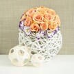 Set on a clean white surface, a beautifully crafted floral display radiates understated elegance, as if prepared by Flowers Stanmore for a wedding breakfast at a nearby country club. The centerpiece is a full, spherical mound of roses, each one a gentle shade of peach, apricot or soft coral. Their petals are lush and layered, pressed closely together so the overall shape appears smooth, like a perfectly rounded cushion of colour. This warm, pastel dome is carefully framed at the base by a circular band of lisianthus flowers. The lisianthus are pristine white in the centre, but their outer petals are traced with a vivid violet-purple edge, adding a crisp, decorative outline that accentuates the ruffled texture. Between these blooms, airy sprays of white baby's breath are lightly threaded, their tiny blossoms scattering subtle highlights and preventing the composition from feeling too dense. All of the flowers are arranged in a distinctive white woven container, bulbous and rounded, with strands of wicker or rattan crisscrossing to form an open, basket-like lattice. Light filters through the gaps, creating soft shadows and giving the piece a sculptural, handcrafted character. Placed in front of the vase are two natural-toned decorative spheres, made from narrow, pale wooden strips intertwined into open orbs. One is larger, one smaller, and together they echo the lattice pattern of the container while adding an informal, organic touch. The softly textured beige background wraps the entire scene in a warm, neutral glow, enhancing the romance of the peach roses and the drama of the purple edges, making this arrangement ideal for intimate celebrations and beautifully dressed tables throughout Stanmore.