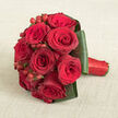 A compact, domed bouquet of luxurious red roses lies diagonally across a pale beige, slightly textured fabric, its richness of colour standing out vividly against the calm background. The roses are densely arranged in a neat, rounded cluster, each bloom brimming with velvety petals in shades of intense crimson and deep claret. Some petals curl gently outward, catching the light, while inner layers remain tightly coiled, giving the bouquet a sense of depth and vitality. Between the roses, clusters of small, round red hypericum berries peek through, their glossy surfaces like tiny drops of lacquered colour, bringing a playful texture to the otherwise smooth composition. Encircling the rose heads, broad, dark green leaves have been intricately folded and wrapped into a tailored collar that holds the flowers together, their slightly glossy finish reflecting light in soft, subtle glints. The stems are bound with a strong, rich red satin ribbon, tightly wrapped yet visually soft, forming a handle that looks ready to be held during wedding vows at a church near Stanmore Hill or in photos at nearby parks. The beige fabric underneath, reminiscent of fine linen or natural cotton, offers a gentle contrast that feels warm and inviting rather than stark. The overall effect is one of elegant romance and thoughtful design, as though the bouquet has just been finished on the workbench at Flowers Stanmore, carefully crafted for delivery to mark an anniversary, engagement, or intimate celebration where every bloom is meant to communicate deep affection.