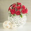 In the centre of a bright, minimalist setting, a dome of red roses and white gypsophila creates a striking yet tasteful display, reminiscent of a carefully styled home near Stanmore Broadway. The bouquet is tightly composed, with about a dozen roses packed closely together into a rounded form. Each rose is a deep scarlet, its petals plush and velvety, curving inward in perfect spirals that suggest peak freshness. Among the roses, soft sprays of baby's breath weave through the blooms, their tiny white flowers clustering in airy bunches that resemble a fine mist or light snowfall, gently balancing the intensity of the crimson. A slim brim of dark green foliage can be seen at the base, providing a subtle boundary between the mass of flowers and the vessel. The arrangement is cradled in an eye-catching, globe-shaped vase, constructed from slender white strips that intersect to form an open, basket-like lattice. This woven sphere has a contemporary yet slightly rustic feel, similar to decorative pieces you might see in a stylish Stanmore living room. The open weave allows glimpses of the inner structure and lets light slip through, casting delicate shadows onto the smooth white tabletop below. On the surface beside the vase, two decorative balls made from pale, natural wicker rest in an informal grouping, their soft cream tones and fibrous texture echoing the craftsmanship of the vase. Behind the scene is a restrained backdrop of pale beige or light grey, with a faint texture that avoids glare or distraction. Soft, even lighting flows over the flowers and their container, enhancing every contour and surface without harsh contrast. The result is an image that radiates measured romance and refined simplicity, well suited to wedding flowers, proposals, or intimate celebrations in and around Stanmore, with same-day delivery from local florists easily imagined.