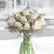The image captures a beautifully balanced bouquet of white roses and lavender statice, displayed in a clear glass cylinder vase resting on a pristine white surface. The roses, pale cream in tone with a whisper of green at the base of a few outer petals, are arranged in a generous, dome-like formation. Each flower's petals spiral inward in precise, layered curves, some tightly furled and others more open, giving the arrangement a gently varied texture while keeping a sense of order. Nestled between the roses are sprays of statice in soft lilac and gentle purple shades, their countless tiny blossoms forming a misty halo that softens the bouquet's silhouette. The glass vase reveals the straight, freshly cut green stems, bundled neatly and immersed in transparent water that glints softly in the light. Natural daylight, similar to the soft light that might filter through a Stanmore bay window, illuminates the scene, casting faint shadows on the white surface and highlighting the roses' velvety texture. The background is out of focus, rendered as a calm blend of pale grey and white, suggesting a modern, uncluttered room somewhere off The Broadway or near Stanmore Hill. The overall feeling is one of gentle dignity and peace, making this design especially suited for sympathy occasions, condolence messages, or quiet remembrance. It is the sort of thoughtful arrangement a Stanmore family might place in their home or send to a service, allowing the serene harmony of white and lavender blooms to express sympathy and support when emotions are tender.