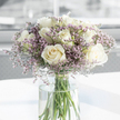 The photograph captures a moment of stillness: a sympathy bouquet of white roses and lilac filler flowers arranged in a clear glass vase, as though placed on a simple table in a Stanmore living room near Brockley Hill. The roses, which form the heart of the arrangement, are fully open and generously layered, with velvety petals curving outward in soft spirals. Their colour ranges from creamy off-white at the centre to a clean, luminous white along the outer petals, creating a sense of quiet sophistication and calm. Interlaced among the roses are slender stems of delicate filler flowers in shades of lavender, pale lilac, and soft pinkish-purple, likely statice. These tiny blossoms cluster together in feathery sprays, adding a misty, pastel veil around the more substantial roses, and lending the bouquet a gentle, consoling character. The flowers are gathered in a tall, cylindrical vase made of clear glass, filled with fresh, transparent water. Through the sides of the vase, the green stems stand clearly in view, neatly trimmed and aligned, underscoring the careful attention of the florist and the freshness of the blooms. The vase rests on a smooth, bright white surface that reflects some of the soft natural light filtering in from an unseen window. The background is softly blurred into pale, neutral tones, suggesting a peaceful, uncluttered interior space somewhere close to Stanmore Country Park, where the bouquet can quietly offer comfort. Together, the roses and lavender fillers create an impression of gentle sympathy, respect, and a tender gesture of support for someone in the Stanmore community during a difficult time.