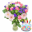 A bright, celebratory tableau: a lush bouquet of fresh cut flowers in a clear glass vase sits against a clean white background, partnered with a cheerful cake-shaped foil balloon that proclaims Happy Birthday. The arrangement is rooted in soft pink romance - dozens of blush to deeper pink roses unfurl their velvety petals, each bloom layered and full, while pale pink Gerbera daisies open like smiling faces with buttery yellow-green centers. Throughout the composition, vivid clusters of small purple filler blooms, likely statice or limonium, give crisp texture and a jewel-like contrast to the pastels. Small creamy white spray flowers tuck into the mix, adding lightness and volume, and glossy deep-green leaves of different shapes frame the bouquet, their smooth surfaces catching the light and making the colours sing. The stems and water are visible through the vase, a simple, honest touch that speaks of freshness and careful handling. To the lower right, a white cake-shaped Mylar balloon decorated with blue piping, multicoloured sprinkles, and a playful border of confetti and stars adds instant fun; four striped candles - purple, red, orange and blue - mark the top, each tipped with a tiny yellow flame, while Happy Birthday is printed in bold multi-coloured letters across the front and a small KALEIDOSCOPE logo sits at the base. Soft daylight seems to lift the scent - a faint, sweet perfume of roses and garden greens - and the whole composition reads as an effortless birthday gift, suitable for delivery around Stanmore, whether picking up on Stanmore High Street or bringing a smile to someone near Stanmore Common.