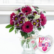 On a smooth, light-coloured surface bathed in soft daylight, a joyful floral arrangement in a clear glass vase is paired with a gleaming heart-shaped balloon, capturing the spirit of a thoughtful gift delivery in Stanmore. The bouquet is full and rounded, anchored by striking gerbera daisies in a bold magenta tone. Their broad, velvety petals radiate around deep, almost black centres, giving the arrangement immediate impact. Intertwined among the gerberas, smaller chrysanthemums crowd together in generous clusters, each floret flaunting deep purple petals with crisp white edges, circling bright yellow centres that bring a lively, almost playful energy. Creamy white roses are dotted throughout, their gently spiralled petals introducing a soft, romantic note that tempers the intensity of the purples and pinks. Airy sprays of tiny white baby's breath weave in and around the other flowers, adding a delicate, cloud-like veil and helping to blend the colours into a harmonious whole. Fresh green foliage peeks out between the blooms and around the edges, contributing both structure and the scent of newly cut greenery. The stems are gathered into a rounded, transparent glass vase filled with clear water, the cut ends neatly aligned to show professional care. Around the vase's neck, a piece of rustic twine is tied into a casual bow, suspending a small white gift tag decorated with a pink rose icon and handwritten-style text, suggesting a personal, locally crafted gesture that might easily come from a Stanmore florist. To the right of the vase, slightly leaning in, a shiny foil balloon shaped like a heart reflects the light from its silver border, while its white centre displays vibrant, patterned letters spelling out an affectionate message and a bright 24-7, underscoring constant hugs and love. With its softly blurred, bright background reminiscent of a window looking out over Stanmore's leafy streets, the whole scene radiates warmth, celebration, and heartfelt intention.