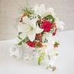 An exquisitely styled floral centrepiece is set on a smooth white tabletop, framed by a softly textured light beige background that recalls the calm, neutral décor of elegant Stanmore reception rooms. At its heart is a round white container fashioned in an open-weave basket style, its lace-like lattice creating a sense of airiness and craftsmanship that feels both contemporary and timeless. From this vessel rise several impressive white oriental lilies, their large, trumpet-like petals fully open and luminous, revealing slender stamens tipped with warm golden pollen. Nestled among the lilies are velvety red roses, mostly in tight bud or early bloom, their rich crimson tones adding passion and depth, ideal for capturing the romance of a Stanmore wedding or vow renewal. Threading delicately throughout are clusters of white baby's breath, each tiny flower contributing to a soft, clouded effect that wraps the stronger shapes in a gentle haze. Small groups of glossy red hypericum berries punctuate the arrangement, bringing hints of festive cheer and added texture. Wispy green foliage, fine and feathery, emerges between the blooms, giving the design lightness and ensuring it never feels too dense. To the right of the main bouquet, a cascade of white phalaenopsis orchids flows elegantly downward, each bloom perfectly formed with subtle yellow and blush-pink centres, while a few buds at the tip remain unopened, suggesting evolving beauty. On the left of the white surface, several white woven spheres rest casually, mirroring the container's texture and balancing the composition. The overall impression is one of refined celebration and romantic poise, perfectly suited for wedding tables, registrar signing areas, or sophisticated events throughout Stanmore, from The Broadway to the quieter residential streets.