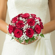 In this elegant bridal image, a round bouquet of rich red roses and delicate white baby's breath is cradled close, as if on the steps of a Stanmore venue just before the ceremony begins. Each rose is beautifully opened, the velvety petals forming layered spirals that catch the light in soft gradients of crimson and shadow. There is a sense of fullness and generosity in the way the blooms press gently together, creating a lush, seamless surface. Threaded between them, sprays of baby's breath introduce a cloud-like softness, thousands of minute white florets that break up the intensity of the red and add a whisper of texture, like fine lace edging a bridal veil. Small glimpses of fresh green leaves add contrast and a feeling of garden freshness, hinting at stems trimmed and prepared in a local Stanmore workshop by experienced hands. The bouquet is shaped into a perfectly rounded dome, its symmetry suggesting careful craftsmanship and an eye for proportion, ideal for holding just below the waist as the bride walks down an aisle in one of Stanmore's churches or function rooms. Against the ivory of the dress, the red roses glow with symbolic meaning-love, admiration, loyalty-while the white gypsophila echoes purity and new beginnings. The imagined fragrance is gentle yet distinct: sweet rose layered over clean greenery, with the soft, powdery note of baby's breath floating in the background. This is not an overly ornate design; rather, it feels classic, refined, and deeply romantic, the sort of bouquet that quietly anchors the emotions of the day and remains timeless in photographs taken around Stanmore Hill or local gardens.