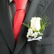 A single ivory rose commands attention here, crafted as a refined boutonniere against the deep charcoal of a formal jacket, each petal soft and slightly open as if breathing. Beneath its luminous center, delicate sprays of baby's breath tumble gently, their tiny white blossoms offering a cloudlike cushion that softens the silhouette and suggests innocence and new beginnings. Narrow fern fronds and trailing glossy leaf strands curve outward, introducing organic rhythm and a subtle verdant energy that keeps the composition feeling alive rather than merely ornamental. The white satin bow tied at the base is small but purposeful, its tidy loops and tails a nod to careful handiwork and ceremonial tradition. Light falls softly across the arrangement - perhaps late-morning light through a Stanmore window near the tube station - highlighting texture and casting mild shadows that emphasise depth and craftsmanship. The contrast between the charcoal fabric and the pale blooms sharpens the rose's tone, making it read as both classic and intimate. There is a faint, clean floral scent suggested by the image, a whispering presence that heightens the sense of occasion. Intended for a groom, best man, or a guest of honour, this boutonniere carries an understated confidence and tenderness. Prepared with local touches familiar to Stanmore residents and often entrusted to florists who know the area around Stanmore Common, it conveys the ritual of marking a special day - small, detailed, and full of personal meaning.
