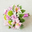 A horizontally styled, hand-tied bouquet rests on a softly textured cream-beige fabric, its colours glowing gently as if caught in late afternoon Stanmore light. At the heart of the arrangement, dusky pink and blush-pink roses unfurl in various stages of bloom, their spiralled centres tightly cupped on the younger buds and more open, ruffled and romantic on the mature flowers. Nestled between the roses are crisp lime-green button chrysanthemums, their neat, spherical heads made up of hundreds of tiny petals that create a playful contrast in both colour and texture. Pale pink and soft peach alstroemeria, with slender petals brushed with yellow and fine burgundy speckles, arc gracefully outward, adding an almost painterly, exotic detail to the bouquet. Threads of delicate white gypsophila weave through the flowers like tiny clouds, softening the transitions and filling any gaps so the composition feels full yet airy. Deep green foliage frames the blossoms, some leaves glossy and structured, others softer and more feathery, guiding the eye along the length of the bouquet. At one end, the natural green stems are gathered together and bound with a vibrant fuchsia satin ribbon, wrapped into a smooth handle that is both practical to hold and visually striking. The ribbon's sheen catches the light, echoing the pink tones of the roses and adding a final flourish of polish. This elegant bouquet, perfect for a Stanmore wedding, anniversary, or special delivery to a home near Stanmore Hill or Canons Park, radiates gentle celebration and timeless romance, while still feeling freshly picked and lovingly arranged.