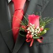 A close-up, high-angle photograph reveals a meticulously crafted wedding boutonniere pinned to the left lapel of a deep charcoal suit jacket, the kind you might see on a groom stepping out by Stanmore Station on a crisp afternoon. At its heart stands a single, velvety rosebud in a rich, almost crimson red, the petals tightly swirled and upright, promising to open as the day unfolds. Around the base of this rose, tiny clusters of white filler flowers, similar to airy baby's breath, form a soft halo, their miniature blooms gently diffusing the intensity of the red. Nestled between them, several glossy, oval red berries catch the light, echoing the rose's hue and adding an almost jewel-like texture. Lush greenery shapes the structure, with slender, grass-like fronds fanning out in elegant spikes and broader, darker green leaves cupping the base of the design with quiet strength. A narrow red satin ribbon, tied into a neat, compact bow, secures the stems and visually links the floral accent to the bright red tie visible beneath the lapel. The tie, smooth and likely satin, is knotted with precision, its vivid colour mirroring the rose and creating a cohesive, confident palette. Beneath, a subtly patterned dress shirt in pale grey and white houndstooth peeks through, adding refined texture without stealing focus. The lighting is soft and even, allowing every curve of petal, shine of berry, and weave of fabric to stand out with clarity. This image conveys the thoughtful detail that goes into men's formal flowers for weddings and proms across Stanmore, where a simple buttonhole becomes a small work of art.